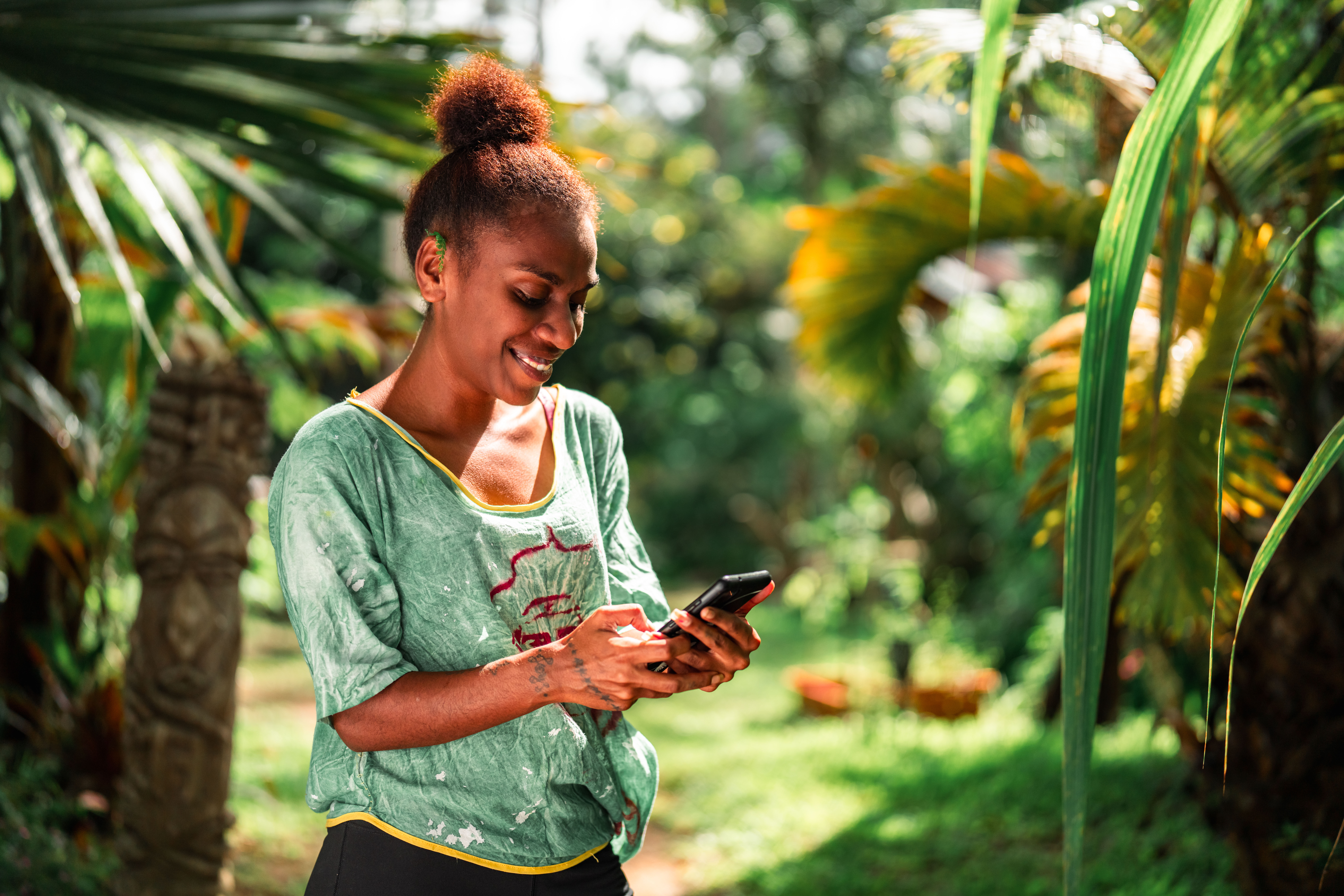Une personne souriante, debout dans un jardin tropical, utilise son smartphone avec concentration, entourée de lumière naturelle et de végétation luxuriante.